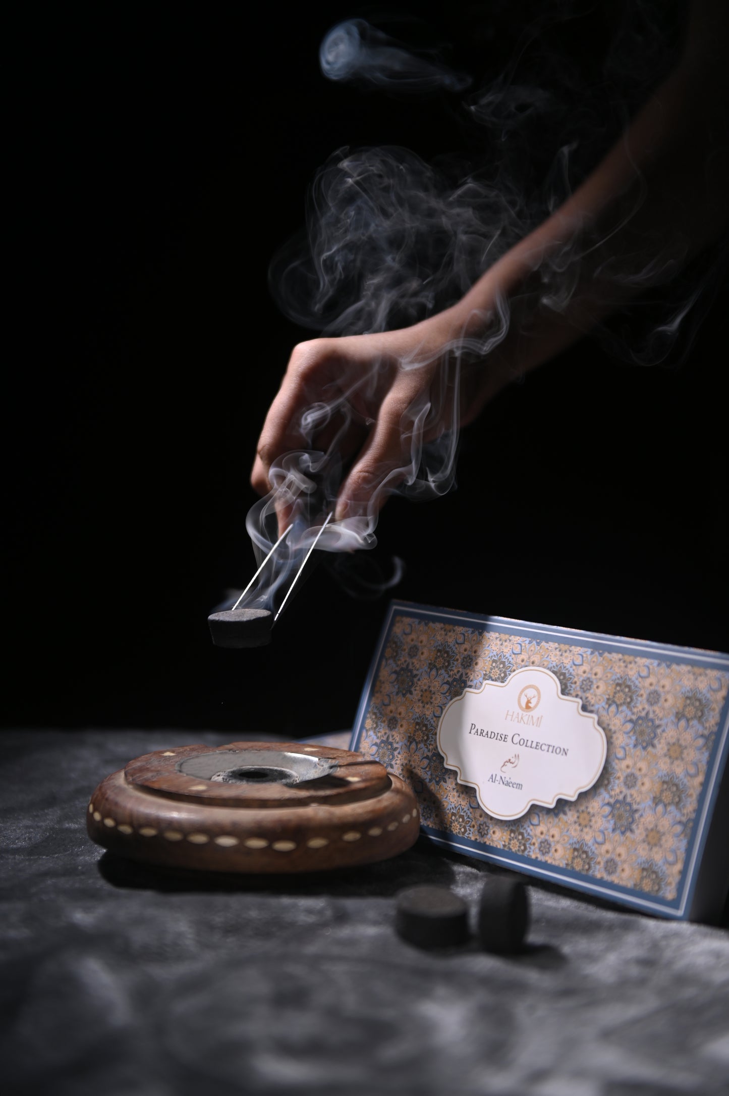 Hand lighting incense with smoke rising, next to an incense burner and box on a dark surface.