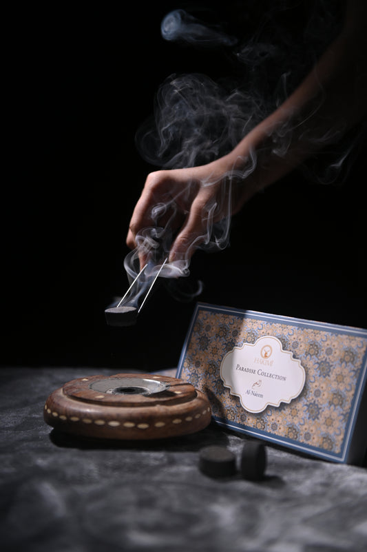 Hand lighting incense with smoke rising, next to an incense burner and box on a dark surface.