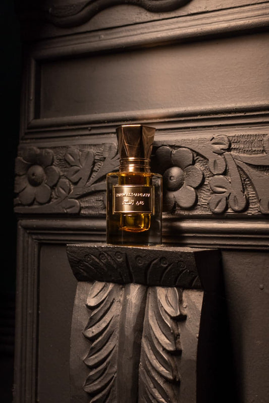 A small rectangular perfume bottle with amber liquid and a metallic gold cap sits on a carved dark stone ledge against an ornate dark wall.