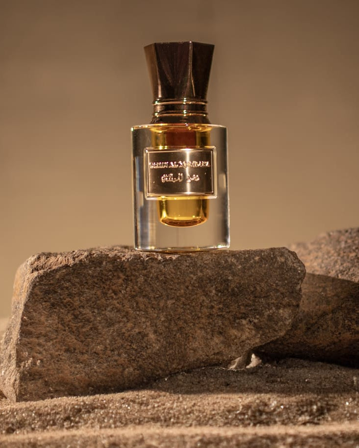 A clear glass perfume bottle with amber liquid and a dark cap, standing on rough stones over sand against a warm beige background. The bottle label reads 'MAISON BOURGEOIS' and 'DES OISIS'.