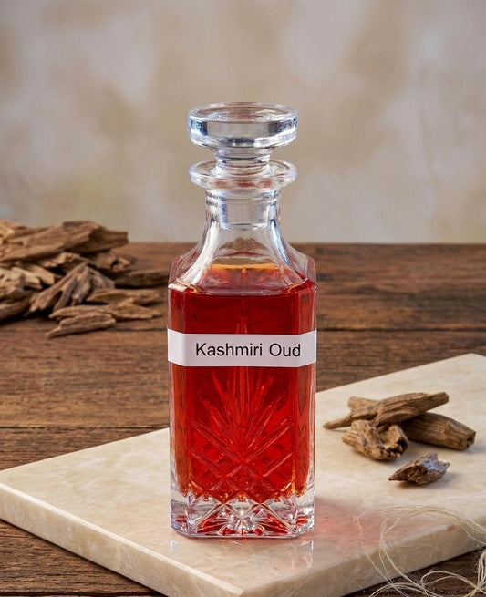 A clear glass decanter filled with red liquid, labeled 'Kashmiri Oud', placed on a light stone slab over a wooden surface with dried botanicals around it.