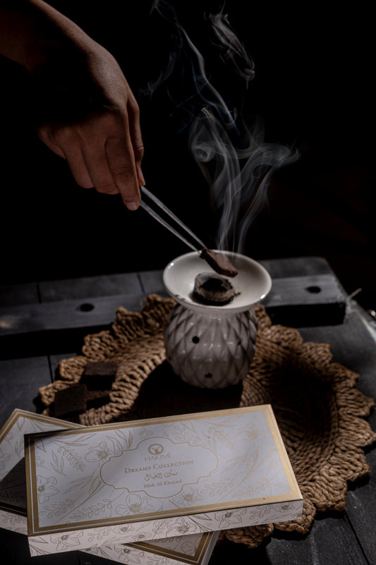 Incense stick being lit with smoke rising from a small incense burner on a textured surface.